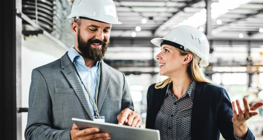 Dois profissionais usando capacetes de segurança brancos conversam dentro de um ambiente industrial. Um deles segura um tablet, enquanto o outro aponta para a tela, indicando análise ou acompanhamento de dados. Ambos vestem roupas corporativas, combinando perfil técnico e gerencial. A imagem representa gestão, colaboração, tecnologia aplicada à indústria e tomada de decisão em campo.