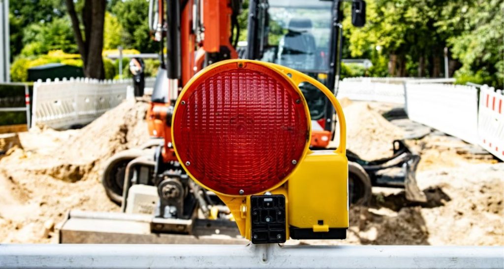 A imagem mostra um sinalizador luminoso vermelho instalado sobre uma barreira de obra, em primeiro plano. Ao fundo, desfocado, aparece uma escavadeira em operação em um canteiro de obras ao ar livre, com terra escavada e estruturas de isolamento. A cena transmite alerta, controle de risco e segurança operacional, típica de ambientes de infraestrutura, obras viárias ou manutenção urbana.