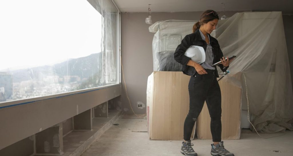 Uma mulher está em um local de construção ou reforma interna. Ela tem cabelo escuro preso e usa óculos de sol na cabeça.