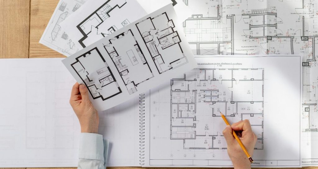 Uma vista de cima (flat lay) de uma mesa de trabalho cheia de plantas arquitetônicas e de engenharia.