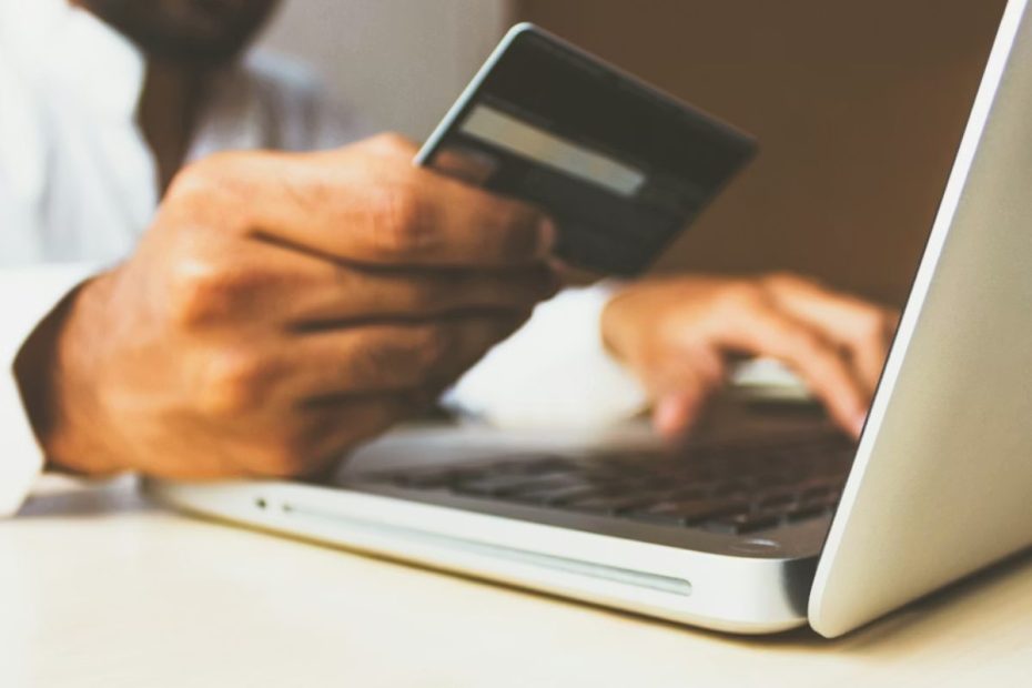 A imagem mostra, em primeiro plano e com foco seletivo (efeito bokeh), uma pessoa utilizando um cartão de crédito ou débito enquanto opera um laptop.