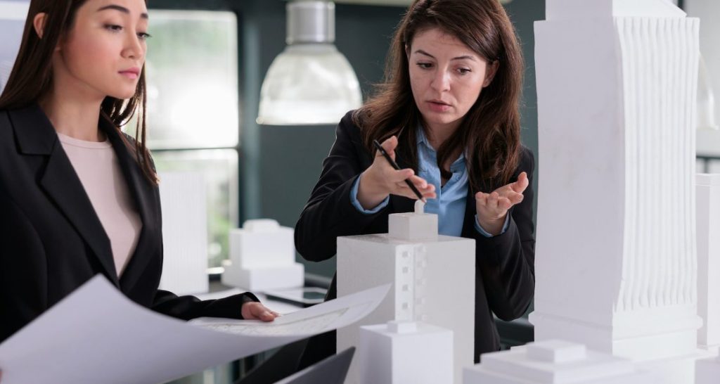 Duas mulheres, ambas vestindo blazers de negócios pretos, estão em um escritório, apresentando ou discutindo um projeto de construção.