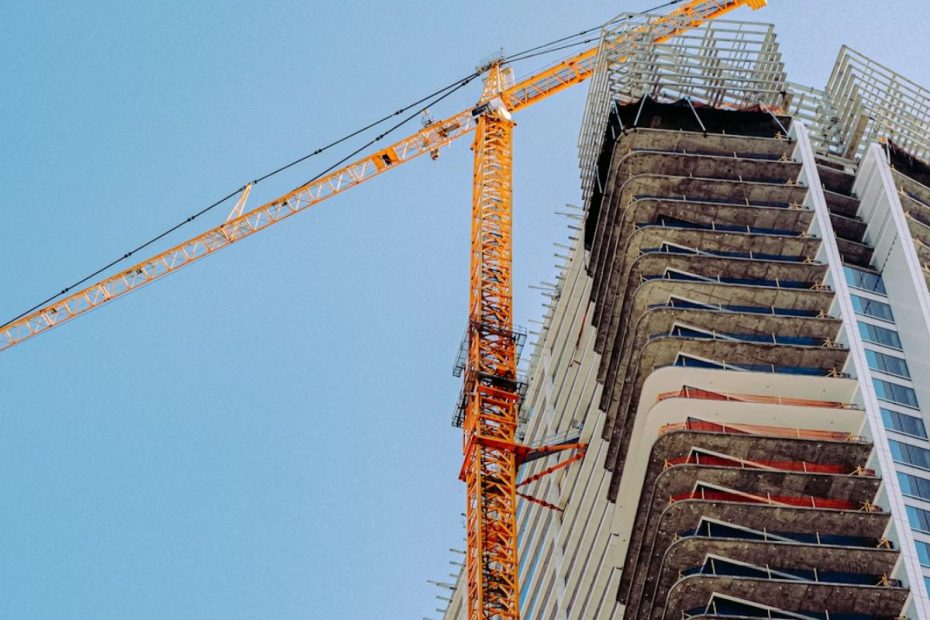 A imagem retrata uma grande grua (guindaste) em um canteiro de obras de arranha-céu, sob um céu claro.