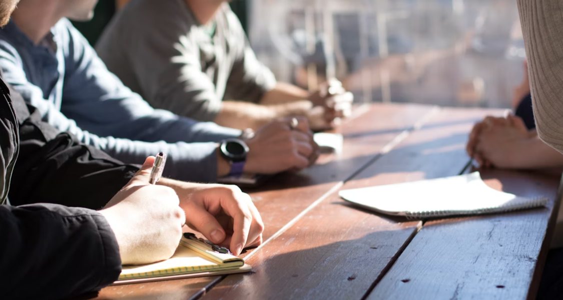 A imagem retrata um grupo de pessoas sentadas em torno de uma mesa rústica, focadas em uma reunião ou discussão.