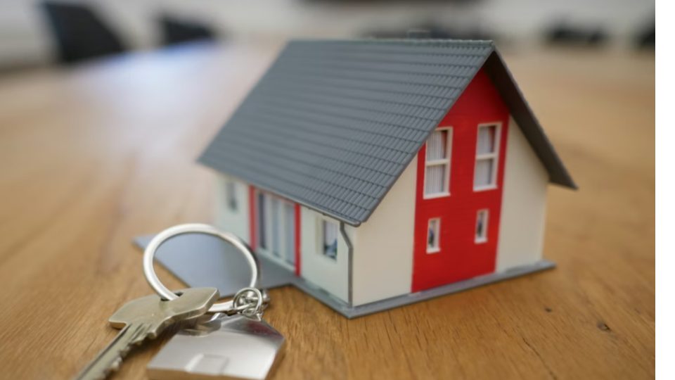 A imagem mostra um modelo em miniatura de uma casa apoiado sobre uma mesa de madeira, ao lado de um chaveiro com chaves metálicas.