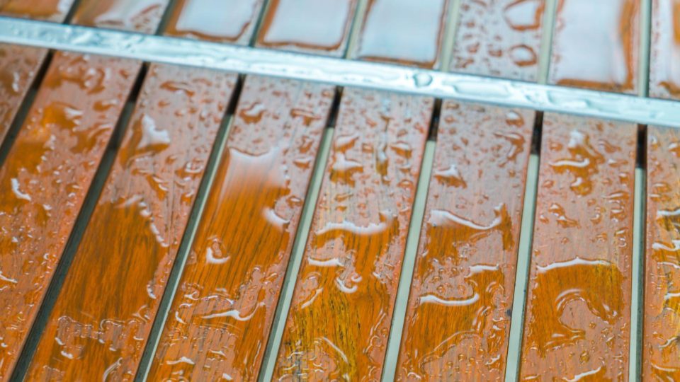 A imagem apresenta um close-up de um piso ou deck de madeira com tábuas alinhadas horizontalmente. A superfície está molhada, com diversas gotas e películas de água espalhadas sobre a madeira, refletindo a luz e destacando o tom quente amadeirado do material.