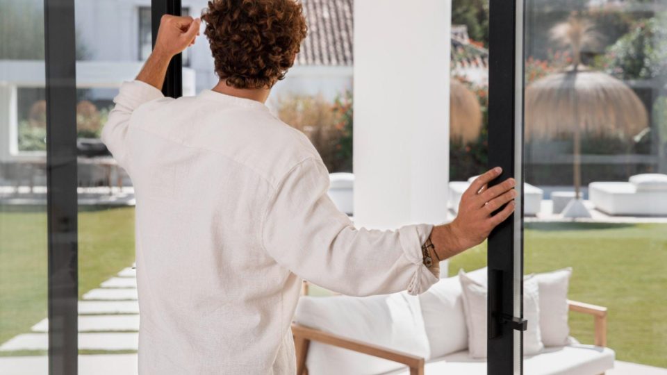 Esta imagem captura um homem de costas, com cabelos cacheados castanhos, interagindo com uma grande porta de correr de vidro em uma residência de alto padrão.