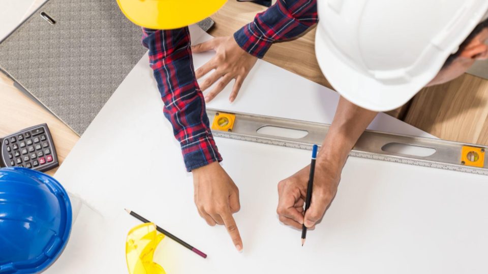 A imagem mostra uma vista superior de dois profissionais da construção ou engenharia trabalhando juntos sobre uma prancheta ou mesa de trabalho. Ambos utilizam capacetes de segurança (um branco e um amarelo), indicando um contexto técnico ou de obra.