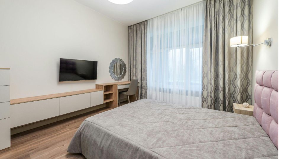 A imagem apresenta um quarto moderno e acolhedor, com decoração clean e paleta de cores suaves. A cama de casal ocupa o primeiro plano, vestida com colcha em tom cinza claro e cabeceira estofada em rosa claro, que adiciona um toque delicado e contemporâneo ao ambiente.