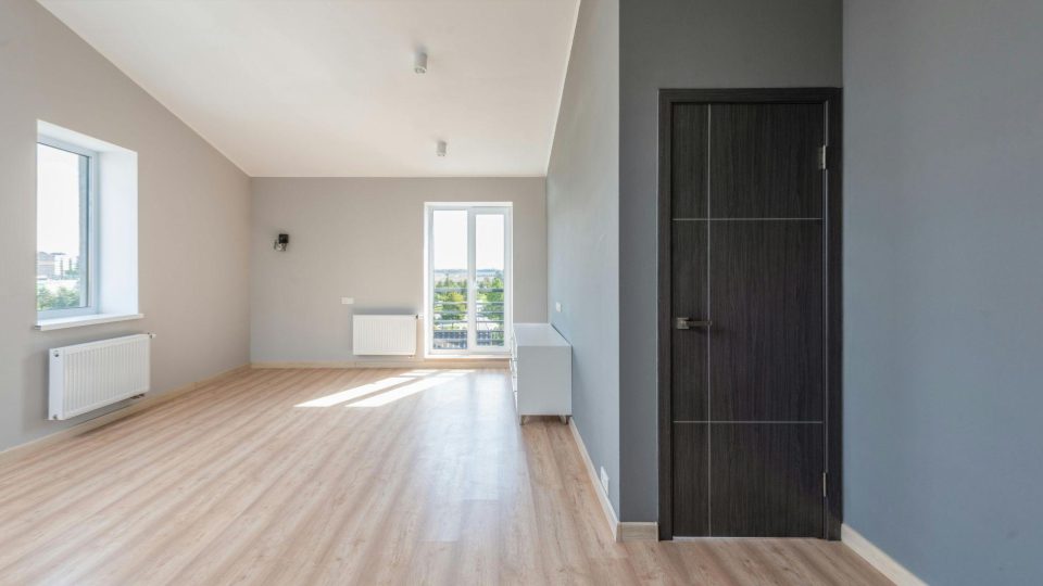 A imagem mostra um ambiente interno amplo e vazio, com design moderno e acabamento minimalista. As paredes são pintadas em tons de cinza claro e médio, criando um contraste sutil e elegante. O piso é de madeira clara, o que contribui para a sensação de amplitude e luminosidade.