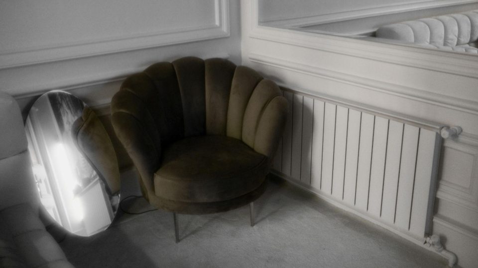 A imagem mostra um canto de ambiente com estética sofisticada e elegante. O destaque é uma poltrona de design arredondado, em tom verde oliva escuro, com estofamento aveludado e encosto em formato de concha, apoiada sobre pés metálicos finos.