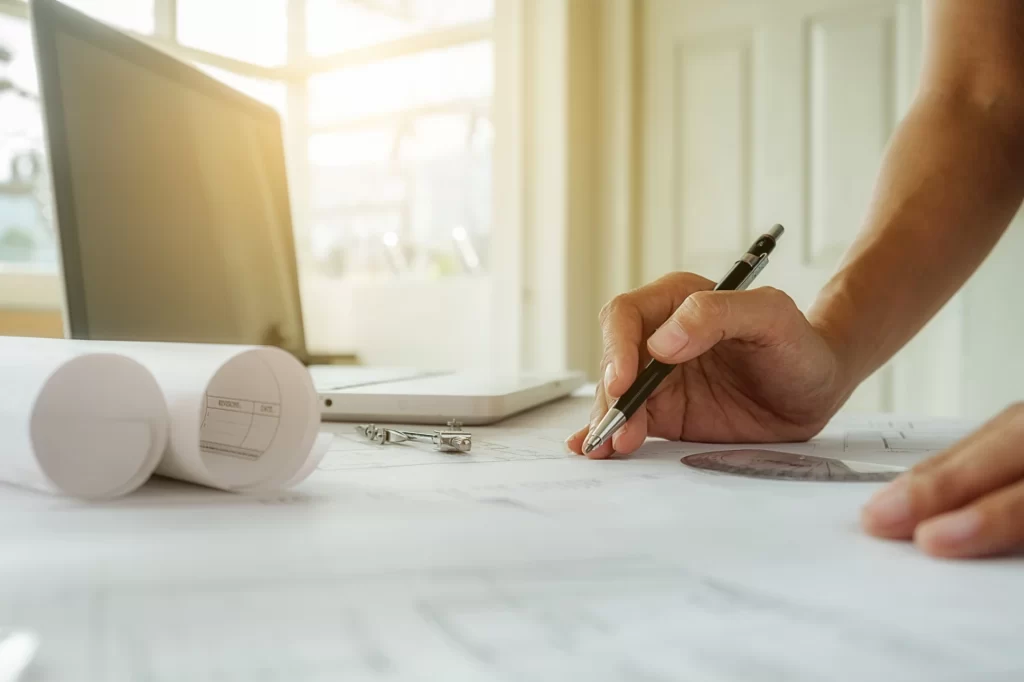 Mãos de um profissional de arquitetura ou engenharia desenhando projetos em uma mesa de trabalho iluminada pela luz natural, com prumo, régua e computador ao fundo