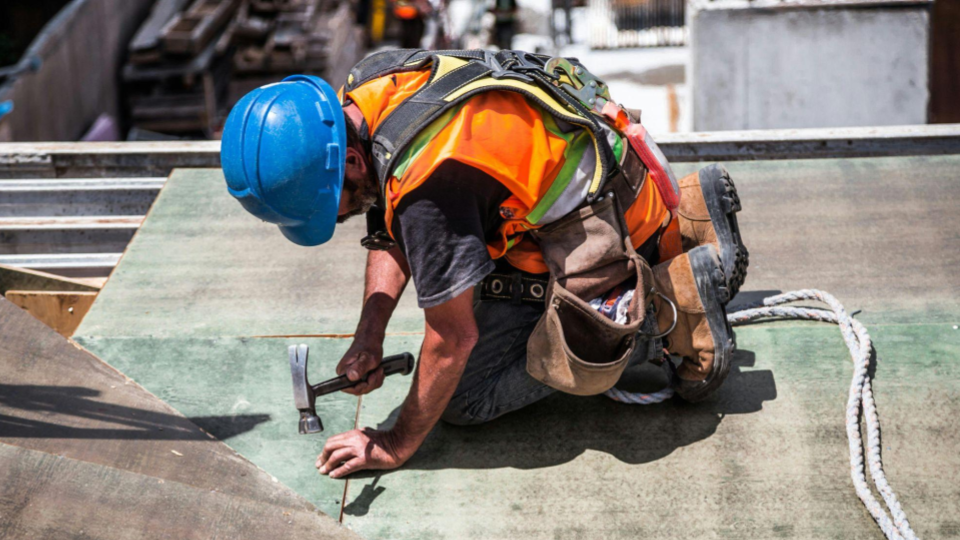 imagem mostra um trabalhador da construção civil ajoelhado sobre uma estrutura metálica ou superfície de obra, utilizando um martelo para fixar um prego ou elemento de fixação.