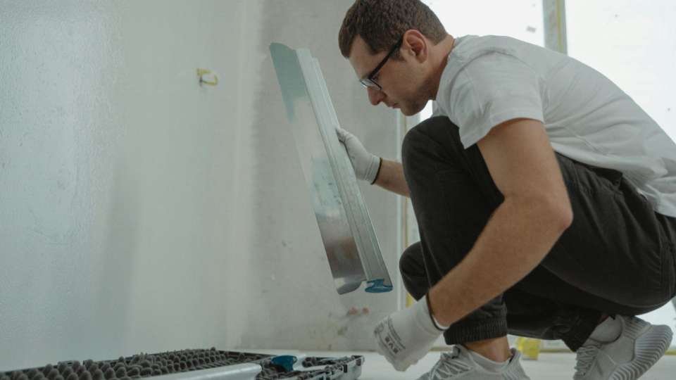 a imagem mostra um homem agachado em um ambiente interno, possivelmente durante uma reforma, segurando uma peça metálica ou placa enquanto trabalha próximo a uma parede.