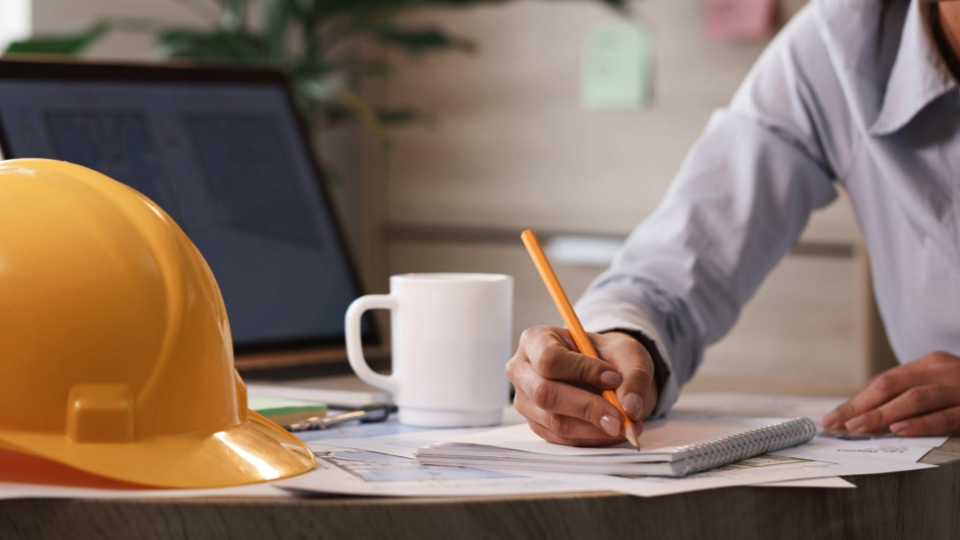 
A imagem retrata um ambiente de trabalho ligado à engenharia ou arquitetura.

Em primeiro plano, à esquerda, há um capacete de segurança amarelo apoiado sobre a mesa, símbolo de construção e segurança no trabalho.