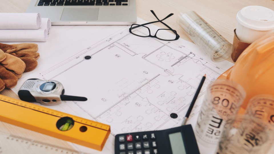 A imagem mostra uma mesa de trabalho organizada com diversos itens ligados à engenharia, arquitetura e construção civil.
No centro, há uma planta arquitetônica aberta, com medidas e divisões de ambientes visíveis.