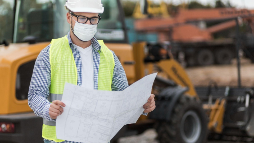 A imagem mostra um profissional da construção civil em um canteiro de obras.

Ele está usando capacete branco, colete refletivo verde, óculos de proteção e máscara facial, indicando preocupação com segurança e normas de proteção.