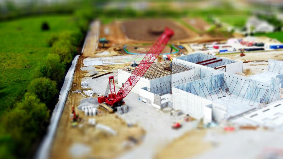 Esta imagem utiliza um efeito fotográfico chamado Tilt-Shift, que faz com que uma cena real pareça uma miniatura ou maquete. Ela oferece uma visão panorâmica de um canteiro de obras em estágio estrutural.