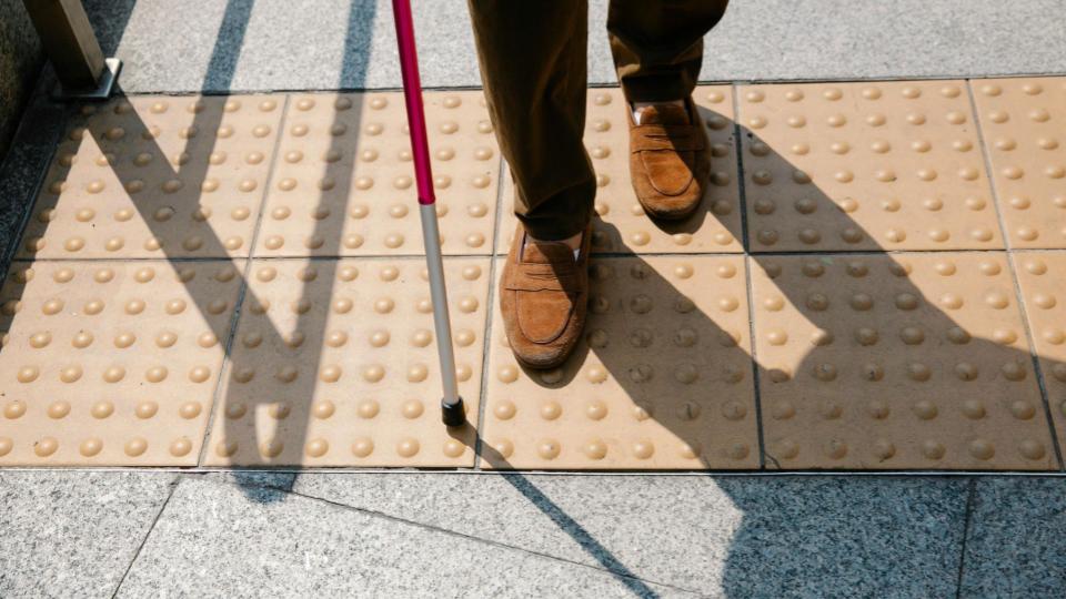 Esta imagem foca nos membros inferiores de uma pessoa caminhando sobre um piso tátil (aqueles blocos amarelos com relevo para orientação).