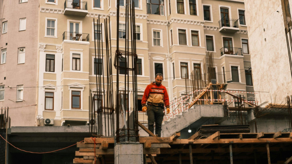 A imagem mostra um trabalhador da construção civil caminhando sobre uma estrutura de obra ainda em fase inicial. Ele veste um moletom vermelho, calça escura e luvas de proteção, olhando para baixo enquanto se desloca com cuidado sobre tábuas de madeira.