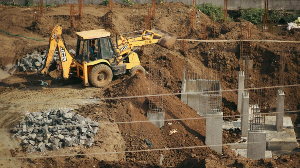 A imagem mostra uma retroescavadeira amarela trabalhando em um canteiro de obras. A máquina está posicionada sobre terra e entulho, com a pá levantando ou movimentando solo.