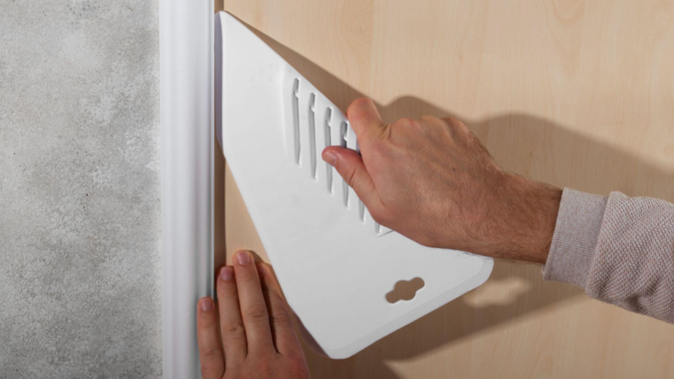 Close-up de uma pessoa instalando um protetor de parede branco (provavelmente de plástico ou borracha) em um painel de madeira. As mãos posicionam a peça cuidadosamente, sugerindo aplicação prática de proteção contra impactos ou desgaste.