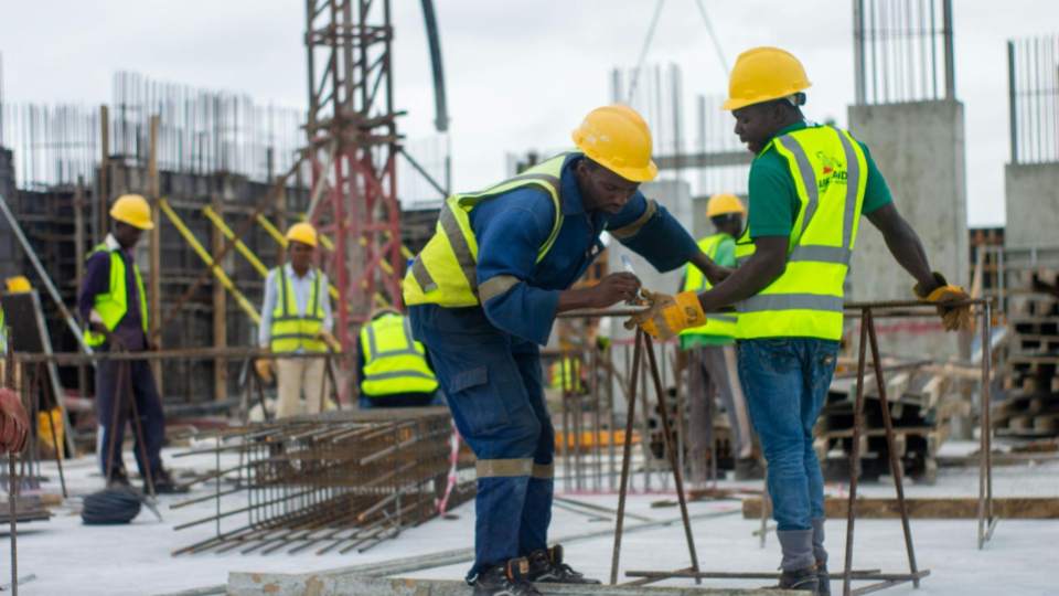 A cena mostra um grupo de trabalhadores da construção civil em atividade. Dois deles estão em destaque, usando capacetes amarelos, coletes refletivos e luvas, enquanto manipulam barras de ferro, provavelmente preparando armações para concreto.