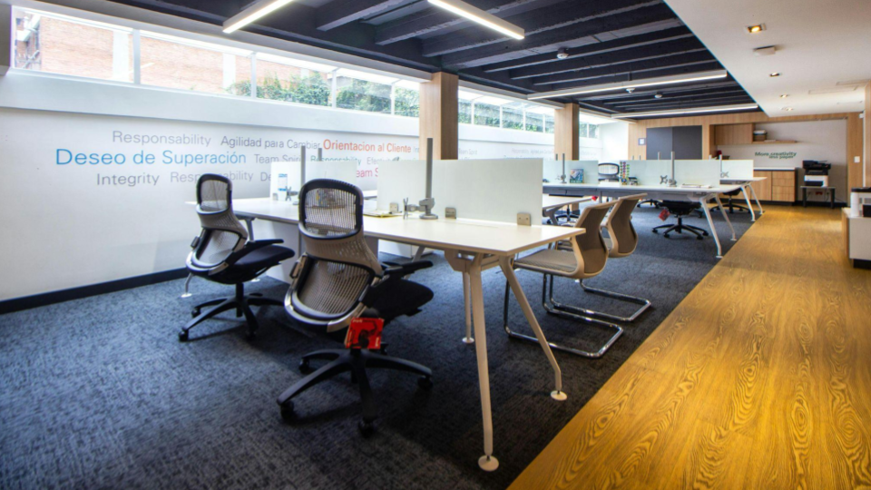 A imagem mostra um escritório moderno em conceito aberto, com um ambiente amplo, organizado e bem iluminado. As estações de trabalho são compostas por mesas brancas compartilhadas, separadas por divisórias baixas, favorecendo a colaboração entre os usuários.