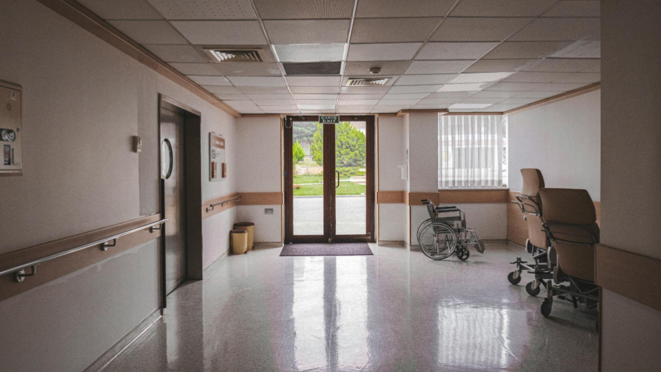 A imagem mostra o interior de um corredor que parece pertencer a um hospital, clínica ou casa de repouso. O ambiente é amplo, limpo e bem iluminado, com piso brilhante que reflete a luz natural que entra pela porta de vidro ao fundo.