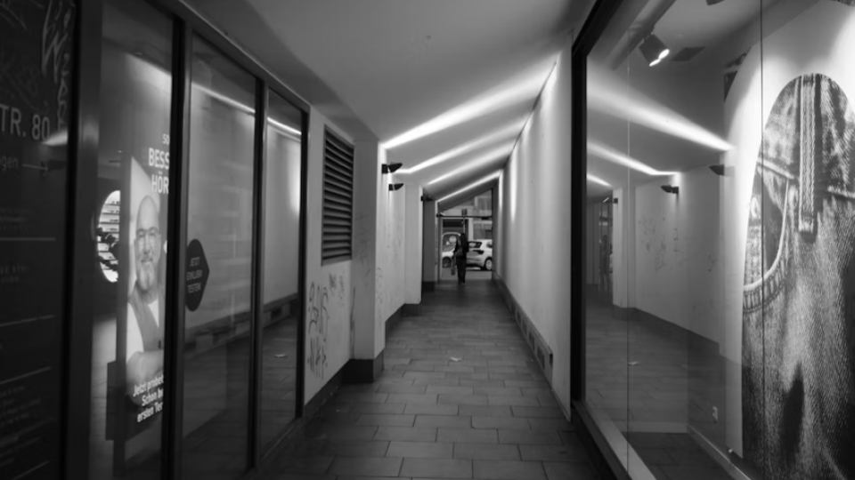 Uma fotografia em preto e branco de um corredor público ou passagem comercial. O destaque é a iluminação linear no teto que cria padrões geométricos nas paredes laterais, com vitrines de lojas ao redor.