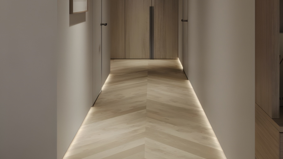 Um corredor minimalista e sofisticado com iluminação de LED instalada no rodapé das paredes, criando um efeito de luz suave no piso de madeira com padrão "espinha de peixe".