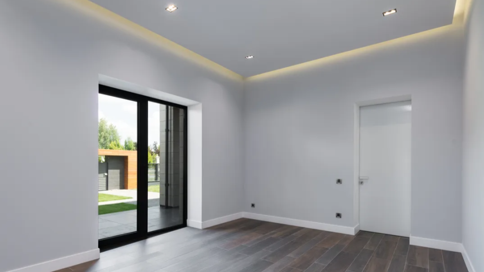 m cômodo moderno e vazio com paredes cinza claro, piso de madeira escura e iluminação embutida (sanca) no teto. Há uma grande porta de vidro preta que dá para uma área externa e uma porta interna branca à direita.