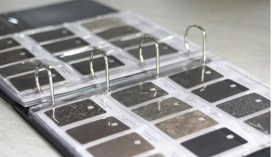 Um mostruário em formato de fichário aberto, exibindo diversas amostras de pedras e granitos em tons escuros e terrosos. Cada amostra possui uma etiqueta de identificação, sendo uma ferramenta típica para seleção de materiais em projetos de arquitetura.