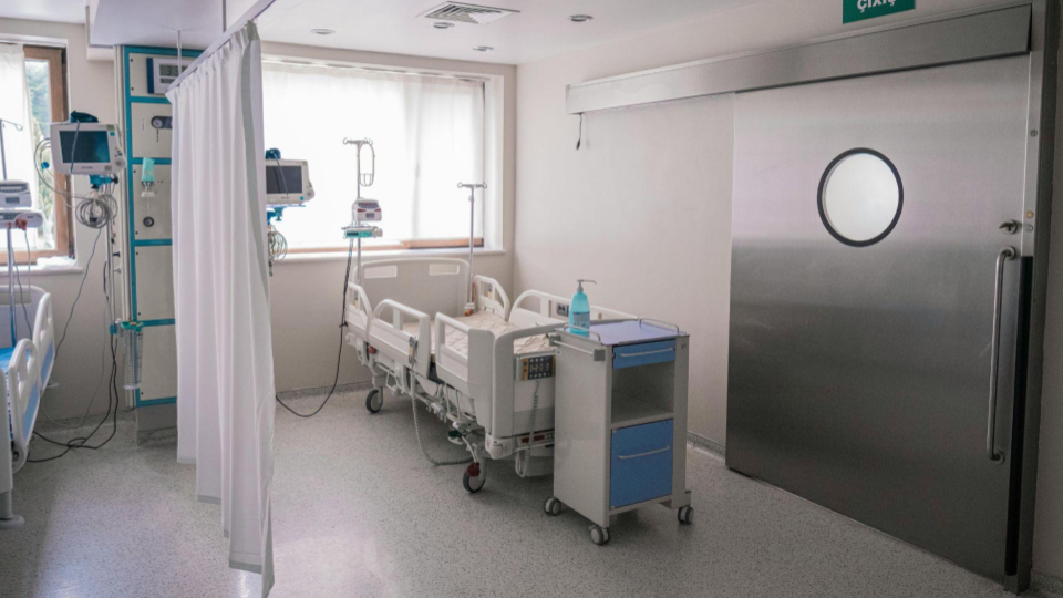 Mostra um leito com equipamentos de monitoramento e uma porta de correr em aço inox. O piso é contínuo, facilitando a assepsia ao redor da maca.