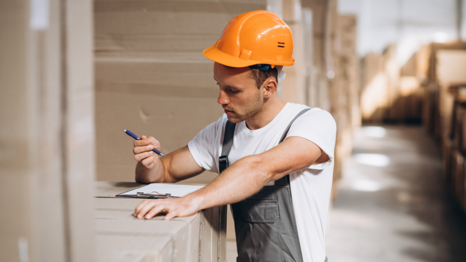 Um profissional utilizando capacete de segurança laranja e uniforme de trabalho faz anotações em uma prancheta dentro de um armazém ou estoque.
