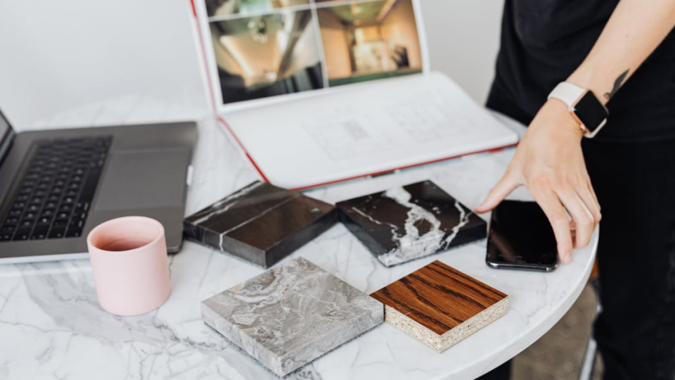 Aqui está uma descrição resumida de cada imagem, seguindo a ordem enviada:

1. Amostras e Planejamento (Mesa)
Uma visão aérea de uma mesa de trabalho com um laptop, uma caneca rosa e diversas amostras de materiais (mármore escuro, pedra cinza e madeira). Uma pessoa manipula um smartphone ao lado das amostras, indicando uma fase de especificação ou escolha de acabamentos.