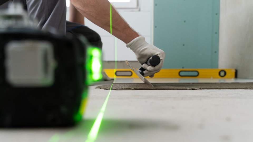 Um close-up de um profissional aplicando argamassa, com foco em uma linha de laser verde que cruza a cena horizontal e verticalmente.