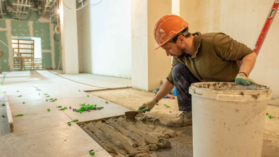 Um pedreiro agachado aplica argamassa no contrapiso usando uma desempenadeira. Ele está espalhando o material em sulcos para garantir a aderência das placas de piso.
