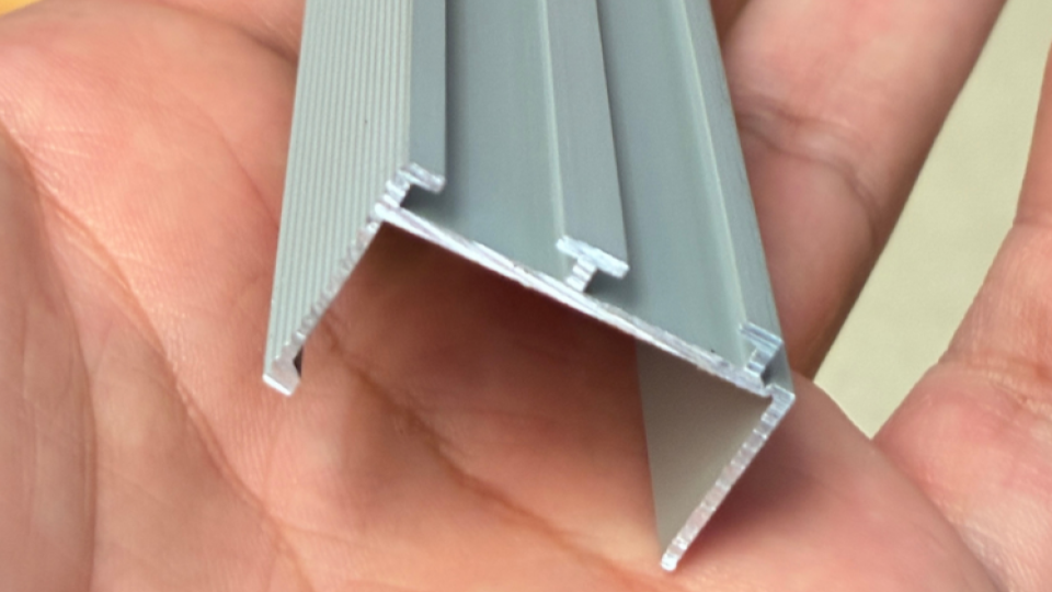 Esta imagem apresenta um detalhe em close-up de um perfil de alumínio extrudado, segurado por uma mão para dar escala.