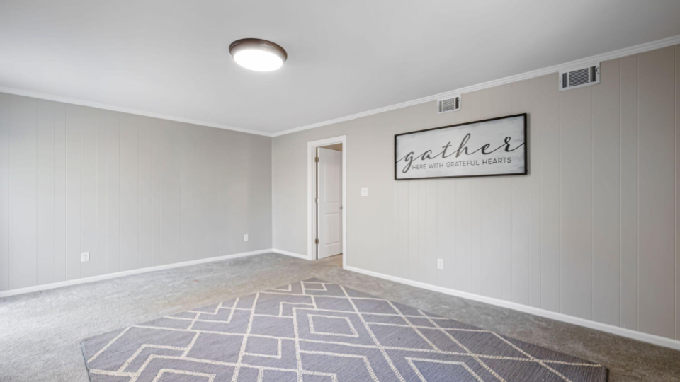 Mostra o interior de um cômodo residencial vazio com carpete cinza e paredes cinza-claro texturizadas com linhas verticais. Há um tapete geométrico cinza e branco no chão e um quadro decorativo com a palavra "gather" na parede.
