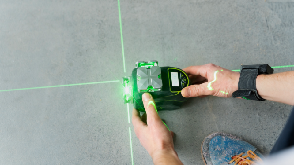 Imagem em plano detalhe mostrando as mãos de um trabalhador operando um nível a laser verde sobre um piso cinza de concreto.