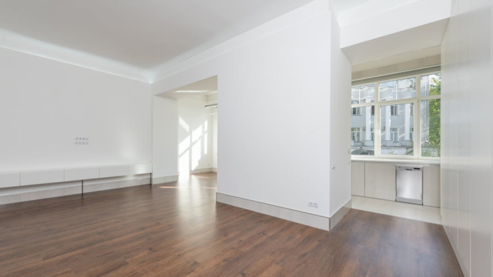 Vista de um apartamento ou casa vazia e ampla, com paredes brancas, piso de madeira escura laminada e uma janela ao fundo que mostra um prédio externo.