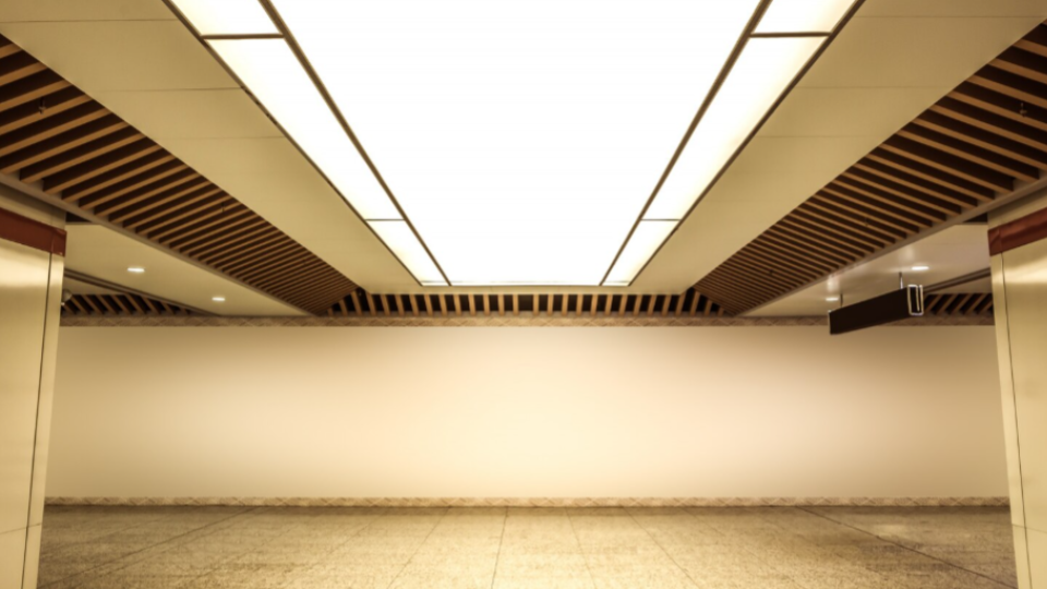 Apresenta a perspectiva central e simétrica de um amplo corredor interno moderno, que lembra uma estação de metrô ou um saguão empresarial. O teto possui um grande painel luminoso central retangular ladeado por estruturas de ripas de madeira inclinadas. O chão é feito de placas de granito claro e o fundo apresenta uma grande parede branca lisa e iluminada.