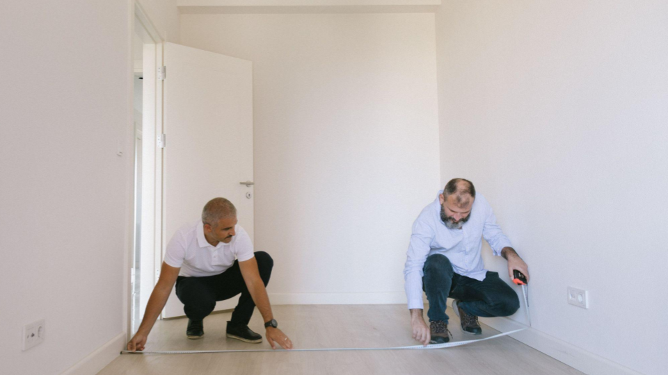 Mostra dois profissionais (possivelmente engenheiros, arquitetos ou instaladores) agachados em um cômodo residencial vazio e pintado de branco. Eles estão trabalhando juntos, esticando uma fita métrica metálica de ponta a ponta ao longo do piso de madeira clara para tirar as medidas exatas do comprimento da parede.