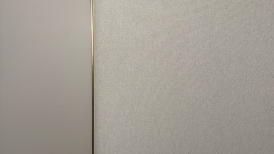 Exibe um plano aproximado e minimalista de uma superfície vertical ou parede dividida em duas texturas. Do lado esquerdo, há uma superfície lisa em tom cinza-claro opaco e, do lado direito, um revestimento texturizado que imita tecido ou papel de parede cinza. Uma fina linha de metal dourado (um perfil ou filete decorativo) faz o acabamento e a transição entre os dois materiais.