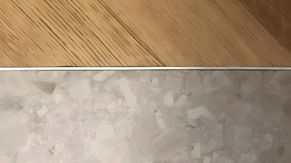 Uma imagem em plano detalhado focada na transição de dois tipos de pisos. Na parte superior, há um piso laminado ou vinílico que imita madeira clara, e na parte inferior, um piso cerâmico ou de porcelanato cinza com textura que lembra granilite ou pedras fragmentadas. Os dois revestimentos são perfeitamente divididos por um perfil metálico (listelo) prateado bem fino.