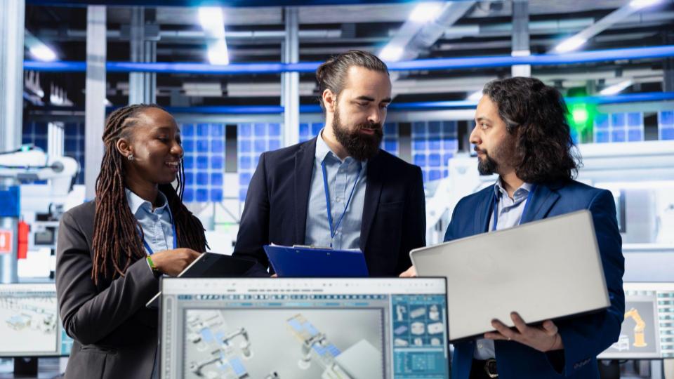 Três engenheiros/especialistas (uma mulher e dois homens) reunidos em uma instalação industrial de alta tecnologia, analisando dados em um notebook, prancheta e monitor.