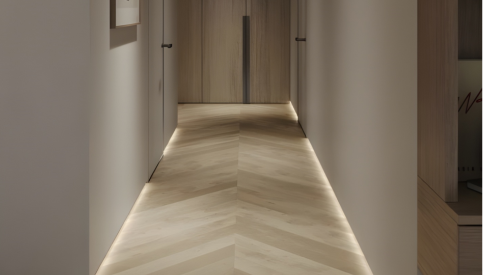 Corredor moderno e minimalista: destaca um piso de madeira com paginação em formato espinha de peixe (chevron) e rodapés invertidos iluminados por fitas de LED, criando uma luz suave e contínua ao longo da base das paredes claras.