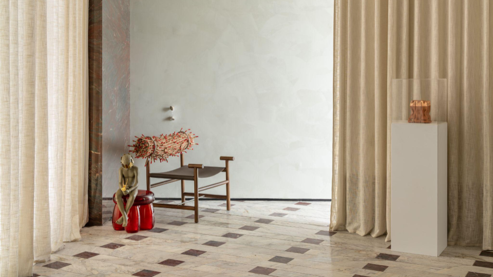 Esta imagem apresenta um corredor amplo e minimalista. Nas paredes laterais, uma textura de cimento queimado na cor cinza claro confere modernidade e neutralidade ao ambiente. O piso de mármore branco com detalhes geométricos pretos adiciona elegância e sofisticação. Ao fundo, as cortinas de linho cru e o mármore avermelhado do balcão anunciam o espaço principal.