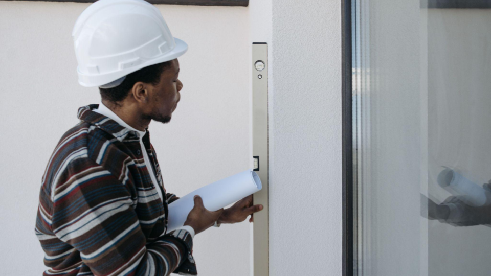 Imagem mostra um homem com capacete de segurança branco e camisa xadrez. Ele segura uma planta arquitetônica e usa um nível de bolha metálico na vertical contra a parede ao lado de uma grande porta de vidro para verificar o alinhamento.